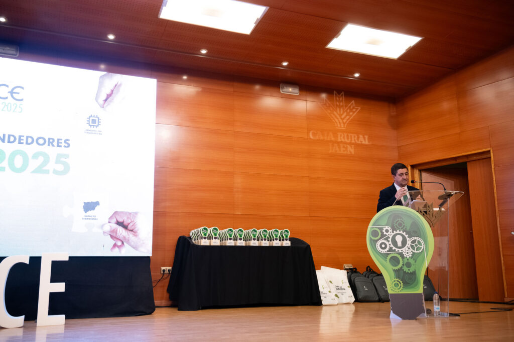 Representante institucional leyendo un discurso en el atril del escenario durante el Foro de Emprendedores Lince 2025, con una mesa llena de trofeos a su izquierda, bolsas y mochilas corporativas al fondo y el logotipo de Caja Rural de Jaén proyectado sobre la pared de madera.