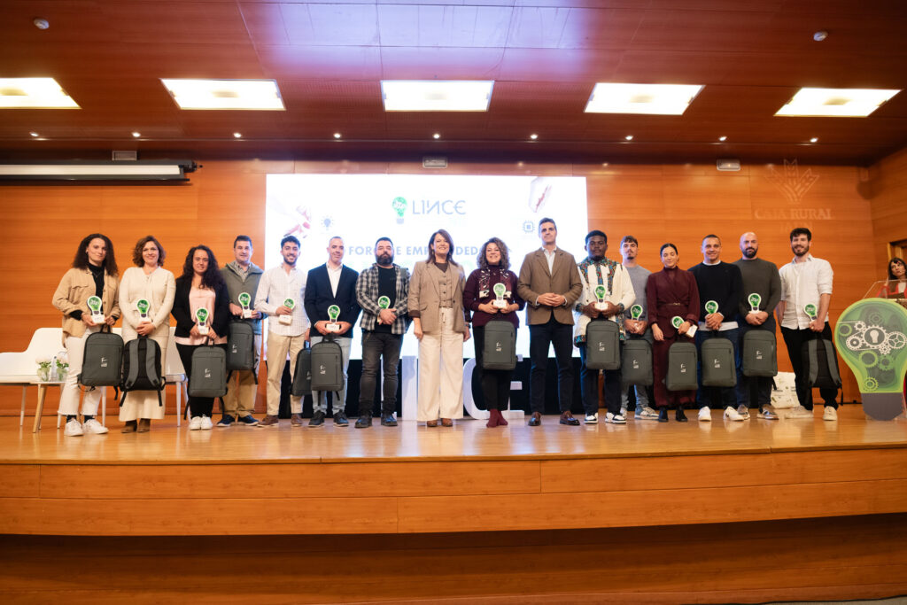 Foto de grupo de los emprendedores premiados en el Foro Lince 2025 posando en el escenario junto a los representantes institucionales, todos alineados frente a la pantalla principal y mostrando sus trofeos con forma de bombilla verde y mochilas corporativas.